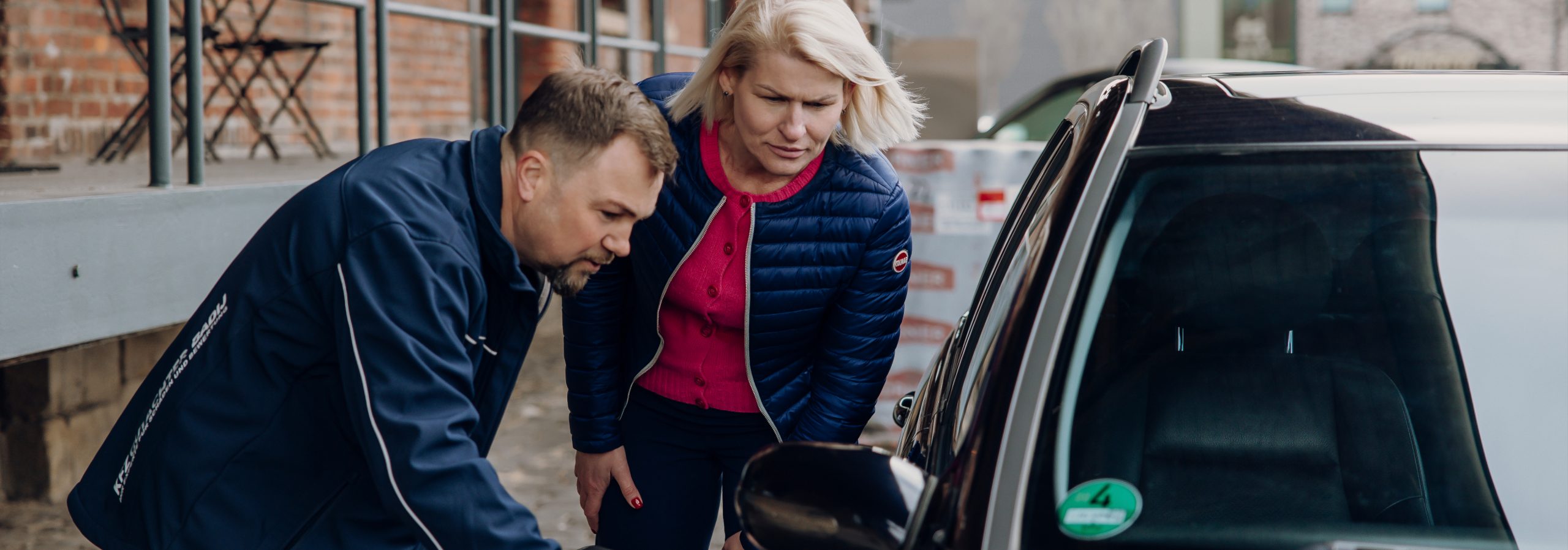 Foto von Viktor Badij mit einer Kundin vor einem Auto. Sie sehen sich den Schaden bzw. Mängel an einem Auto an.